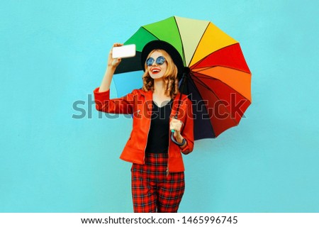 Happy smiling woman taking selfie picture by phone with colorful umbrella in red jacket, black hat on blue wall background