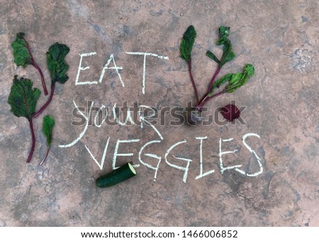 The words eat your veggies written with sidewalk chalk  outside surrounded by fresh vegetables cucumbers and beets