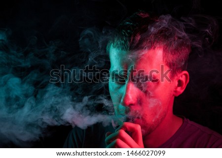 A young brunet male turned his head to the side, holds in his hand and inhales e-cigarette with a vape while smoking and releases smoke to the side, his face is highlighted with red and green light
