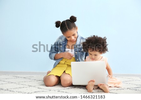 Cute African-American sisters with laptop watching cartoons near color wall