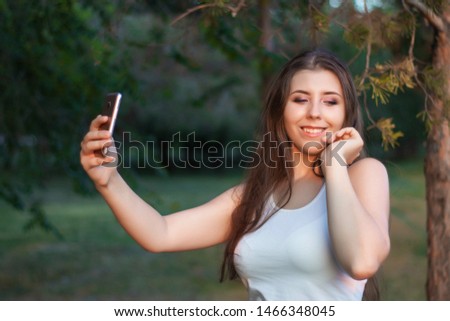 A young woman in a summer park holds a smartphone in her hands, communicates in social networks, takes pictures of herself