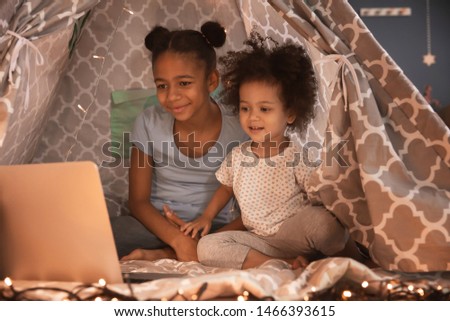 Cute African-American sisters with laptop watching cartoons in hovel