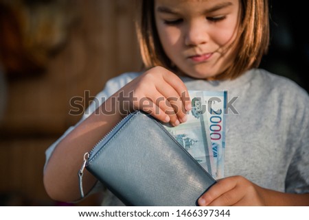 a girl of 7 years of European appearance in a gray T-shirt is holding a leather mother's wallet with Russian 1000 and 2000 rubles bills Royalty-Free Stock Photo #1466397344
