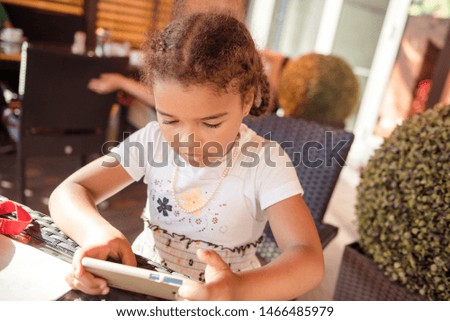 Beautiful little girl playing game or watching video on smartphone mobile. Girl watching cartoons or browsing internet, copy space. Side view portrait of girl using smartphone while sitting.