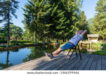 A young man meets the dawn while sitting on a wooden folding chair with legs crossed and holding his palms on the back of his head