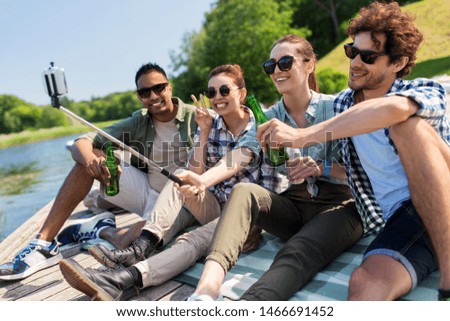 leisure, picnic and technology concept - friends with drinks taking picture by selfie stick on lake pier in summer park