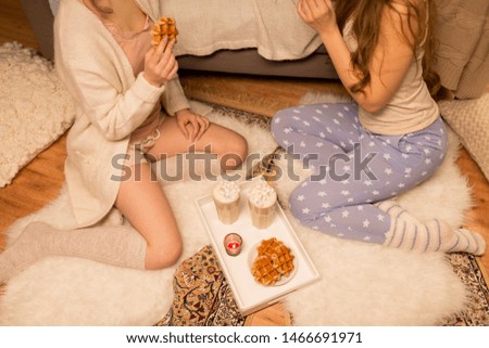 friendship, hygge and pajama party concept - two female friends or teenage girls drinking hot chocolate with marshmallow and eating waffles at home