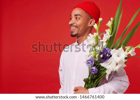 Portrait of young happy African American attractive guy, wears in white shirt and red hat, looks away and holds bouquet , stands over red background and smiling.