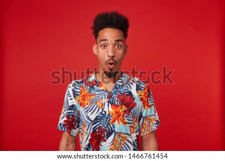 Portrait of shocked young African American guy, wears in Hawaiian shirt, looks at the camera with surprised expression with wide open mouth and eyes, stands over red background.