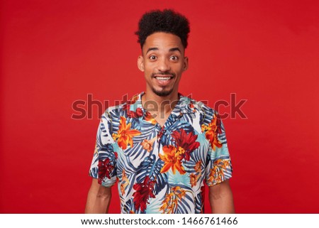 Portrait of happy young African American guy, wears in Hawaiian shirt, looks at the camera with cheerful expression, stands over red background and broadly smiles.