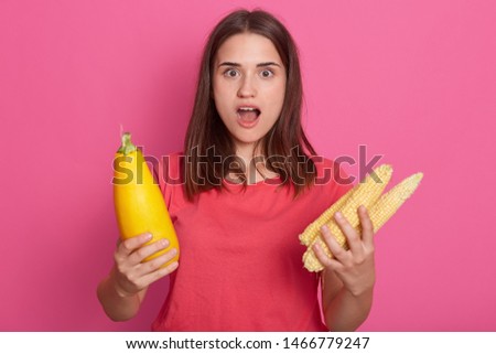 Horizontal shot of young brunette woman holding zucchini and corncob, posing with open mouth. Healthy eating, lifestyle and raw food diet concept, photo isolated isolated over rose background.