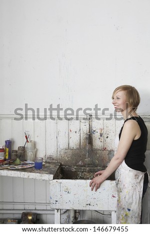Side view of a smiling female artist standing by dirty sink in studio