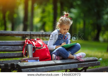 Little beautiful school girl drawing with colored pencils, sitting on a bench in sunny park