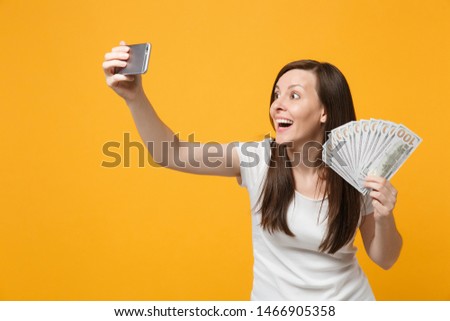 Excited young woman in white clothes doing selfie shot on mobile phone holding fan of cash money in dollar banknotes isolated on yellow orange background. People lifestyle concept. Mock up copy space