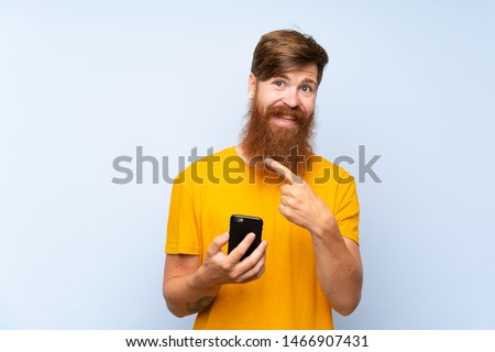 Redhead man with long beard with a mobile over isolated blue wall pointing to the side to present a product
