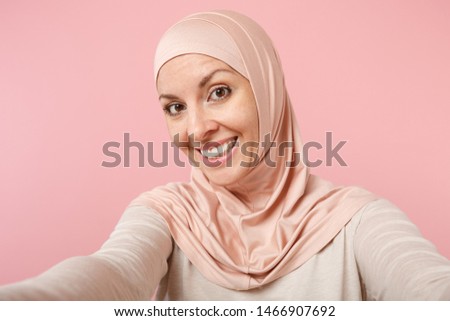 Close up smiling young arabian muslim woman in hijab light clothes posing isolated on pink background. People religious Islam lifestyle concept. Mock up copy space. Doing selfie shot on mobile phone