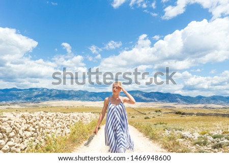 Caucasian young woman in summer dress holding bouquet of lavender flowers while walking outdoor through dry rocky Mediterranean Croatian coast lanscape on Pag island in summertime.