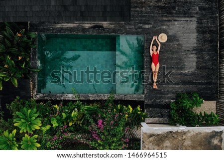 Aerial drone photo of happy Woman in red swimsuit relaxing near private pool with flowers and greenery around, Bali. Tropical background and travel concept. Royalty-Free Stock Photo #1466964515