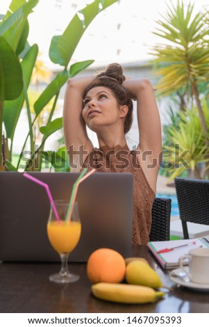 Colorful portrait of a young woman stretching out her arms on the terrace - Fruits, orange juice and her computer on the table - Green background plants - Vertical image