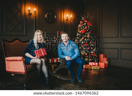 Happy couple sitting by a Christmas tree in a cozy dark living room on Christmas eve. Happy young people give each other gifts by the fireplace near the Christmas tree.