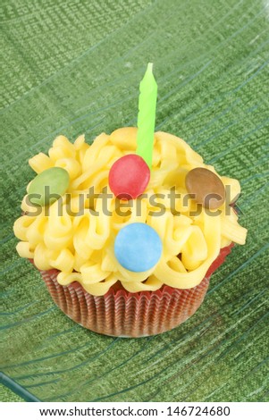 Fancy birthday cupcake decorated with small colorful sugar-coated chocolate confectionery and a green candle on a transparent glass dish, over a green background. Selective focus.