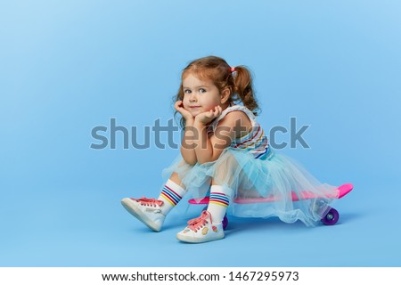 Cool little toddler girl in bright clothes sits on a skateboard and looking at the camera isolated on blue background Royalty-Free Stock Photo #1467295973