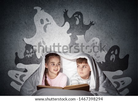 Scared girl and boy reading kids book in bed. Frightened children lying under blanket together. Kids in pajamas and funny ghosts silhouettes back on grey wall. Children reading magic fairy tales