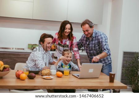 Two charismatic boys with their mature parents taking breakfast together , dad shows something on his laptop and they all together looking , modern kitchen design on the background