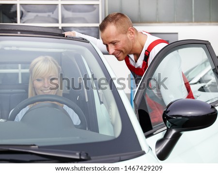 a blonde women sit in her repaired car in front of the car service workshop garage and talk to the professional technician employee in a red overall and looks happy, attractive and satisfied
