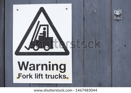 Old Warning Fork lift trucks sign black text and symbol in a triangle with faded background screwed on dark wooden fence.