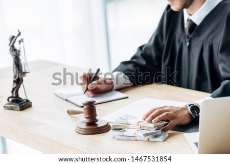cropped view of judge holding envelope with money near gavel and statuette of justice 