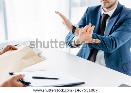 cropped view of businessman gesturing near business partner giving envelope with bribe and clipboard with contract in office 