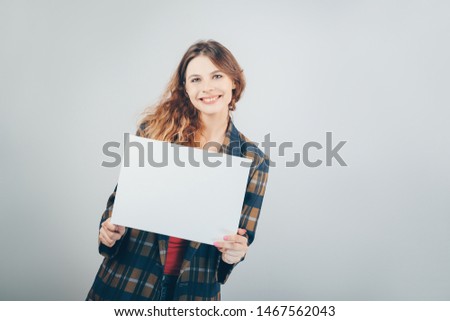 Caucasian girl holding a blank canvas in her hands and smiling, grey wall on background. Copy space. Beautiful young businesswoman with mockup poster.