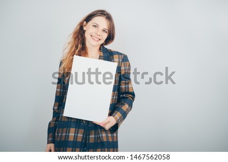 Beautiful girl holding a blank canvas and smiling, grey wall on background. Copy space. Pretty young woman with vertical mockup poster.
