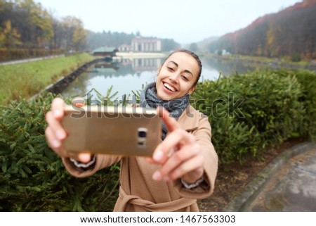 Smiling woman taking a selfie on mobile smart phone camera in the autumn park with lake on background. Beautiful young stylish female wearing long beige coat enjoying view over the lake at cloudy