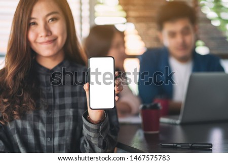 Mockup image of a woman holding and showing black mobile phone with blank screen with colleaques working on laptop in background