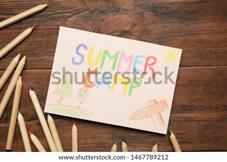 Card with text SUMMER CAMP, drawings and colorful pencils on wooden table, flat lay