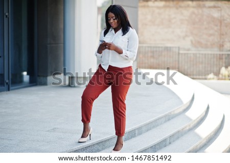 Formally dressed african american business woman in white blouse and red trousers with mobile phone. Successful dark skinned businesswoman.