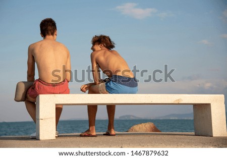 Two young men are sitting with their backs to the camera on a stone bench against the sky and the sea.
