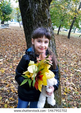 A girl stands near a tree holding in her hand autumn leaves of trees and a teddy bear.
