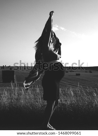 Pregnant woman with beautiful curves during sunset at the edge of the field, shortly after harvest, wrapped in sunlight and summer wind. The girl has a raised hand and holds a scarf in it.