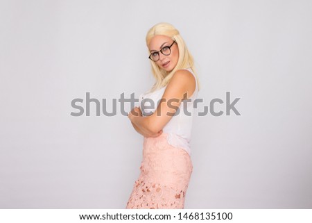 Young blond woman in glasses posing on a white background in the studio. She is wearing a pink skirt and a white blouse.