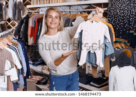 Cheerful attractive girl holding and demonstrating baby clothes on hanger in kids clothing showroom
