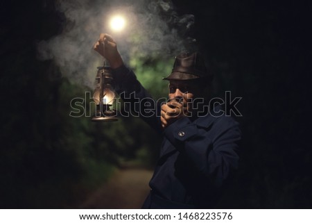 Detective agent in a hat and coat is holding in hand a kerosene lamp and smokes his smoking pipe.
