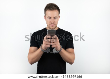handsome young man in black t-shirt holding smartphone while being handcuffed on isolated white background