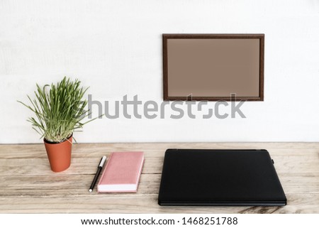 Empty frame on white wall. Closed laptop, notebook and pen. Green houseplant. Home workplace. Place for text