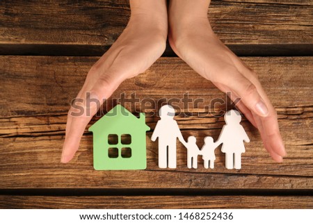 Woman holding hands near figures of house and family on wooden background, top view