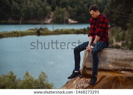 hipster guy in a checkered red shirt sits on a rock on a background of forest and river