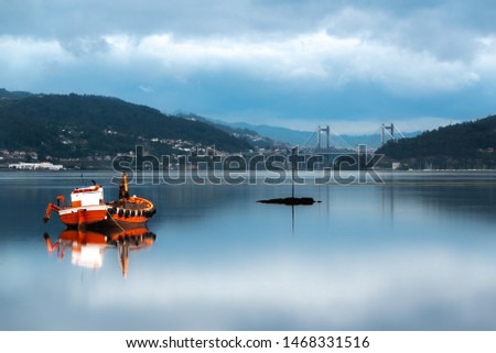 Red fishing boat stranded on water with mountains and one suspension bridge in background. Long exposure photography with reflections in the water, Ría de Vigo, Rande bridge, Vigo, Pontevedra,Galicia.