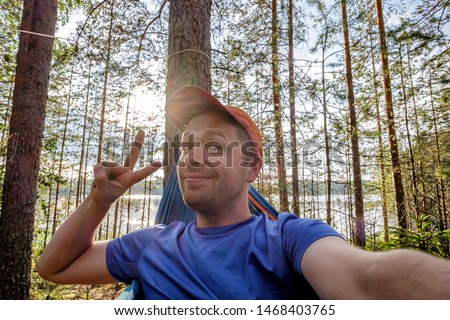 Young man by the lake hanging on hammock relaxing in the morning takes a selfie portrait.